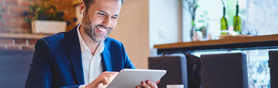 A man in a blazer smiles while using a tablet at a cozy cafe. He is seated near a table with plants and bottles, radiating a relaxed and positive mood.